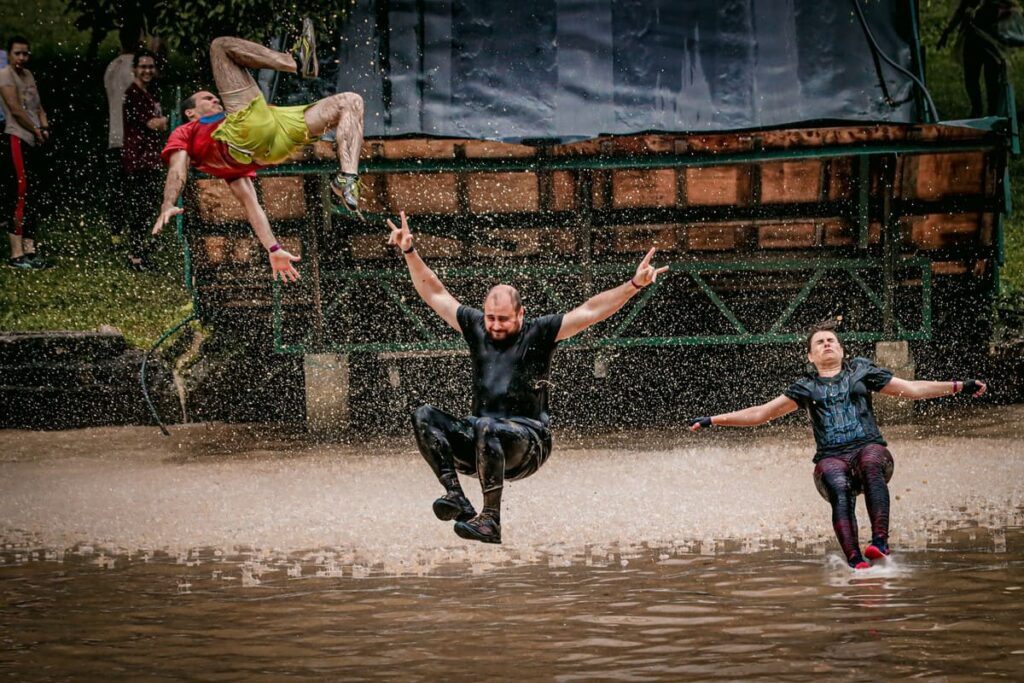 Três corredores molhados pulam em um fosso de água durante a Desafio Braves Mud Race, uma corrida em São José dos Pinhais, exibindo expressões de alegria e determinação.
